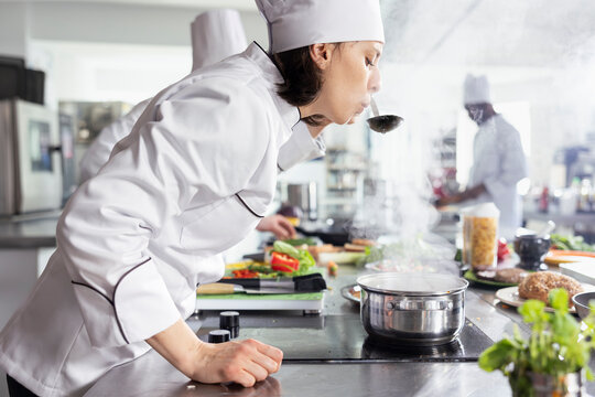 In a busy restaurant kitchen, chef tastes a steaming pan of soup, ensuring perfect seasoning of a great gourmet dish. Culinary workspace stocked with kitchen tools and fresh ingredients.