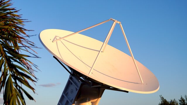 Large parabolic antenna pointing at the sky during sunset, with some vegetation in the foreground, used for aerospace communication and radar applications