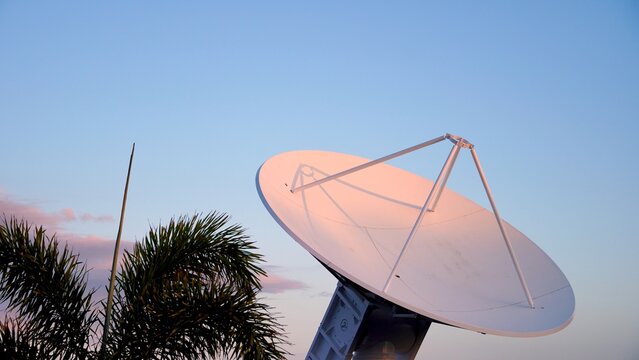 Large parabolic antenna pointing skyward at sunset, framed by palm trees, showcasing warm hues and highlighting advanced technology in space exploration and communication