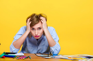 stressed woman sitting at desk surrounded by scattered supplies on yellow background. overwhelmed creative process, frustration in workspace, emotional challenges in professional setting