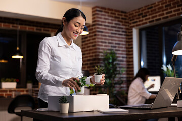 Happy female employee refreshing her workstation, removing plant pots from box and arranging them on her desk. Asian businesswoman standing and smiling, enjoying the process of decorating her office.