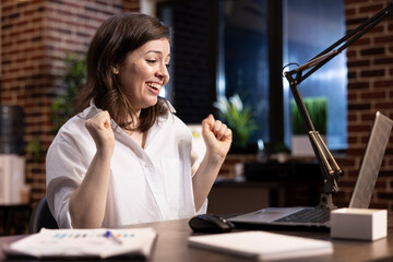 Caucasian female manager in an office receives great news, beaming with excitement as she lifts her arms in joy. She looks at her laptop, feeling victorious and thrilled about her accomplishment.