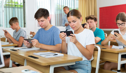 Group of modern teenagers sitting with mobile phones on lesson in classroom