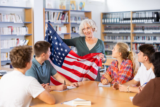 Mature female teacher shows the flag of United States of America to teenagers and talks about this country during lesson at a library school - Powered by Adobe
