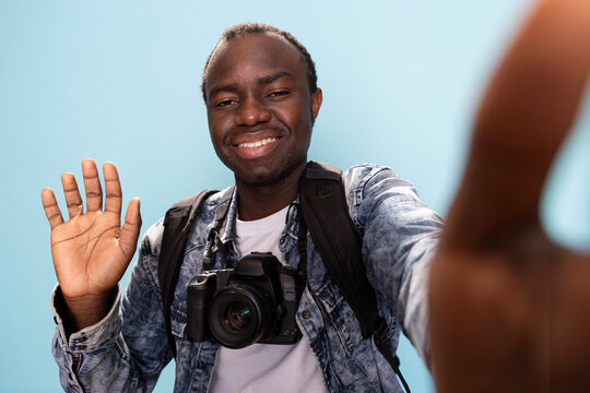 African American man with DSLR around his neck, makes hello gesture, recording a video for his vlogging channel. Black male photographer smiles and waves at the camera in studio with blue background.