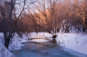 A small frozen river with blue ice in a snow-covered park with sunset sun highlighting tops of bare trees