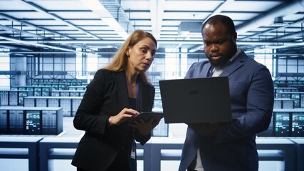 Teamworking colleagues in server farm using laptop to patch network issues affecting transfer rates. Coworkers in data center doing brainstorming, fixing errors affecting supercomputers, camera B