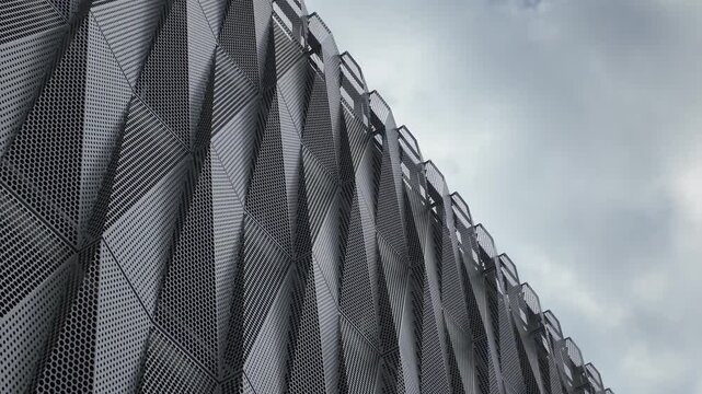 Modern architectural facade featuring dynamic geometric patterns formed by angled, perforated metal panels under a cloudy sky.