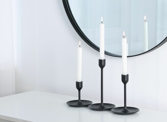 Stylish candlesticks with burning candles on light table near white wall and mirror indoors, space...