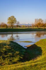 Spring landscape with reflection of trees in the river and snow-capped mountains in the background.