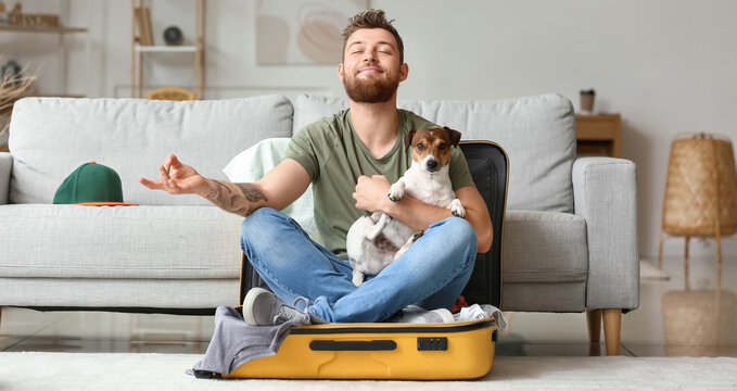 Young man with cute dog meditating in suitcase at home