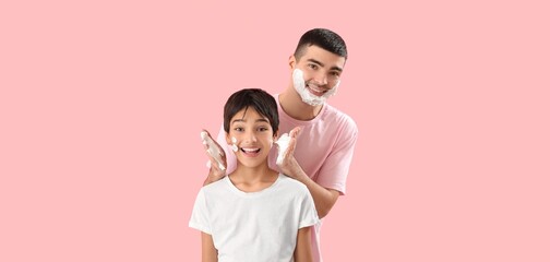 Man and his little son applying shaving foam onto faces against pink background