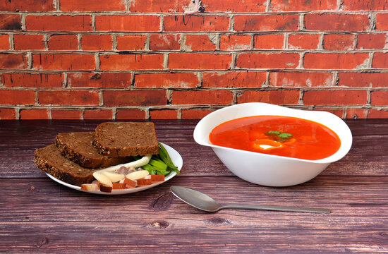 Freshly cooked borscht, tablespoon and plate with pieces of rye bread, garlic, lard and green onions on a wooden table.