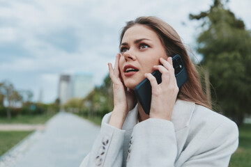 Worried young woman in white blazer speaking on phone outdoors expressing stress and anxiety, urban background with green trees and cloudy sky