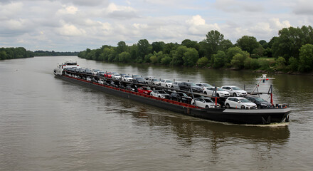 Obraz premium Black River Barge Carrying Numerous New White Automobiles Under Cloudy Skies