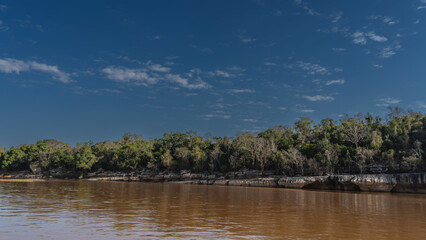 A calm red-brown African river. There are thickets of green trees on the eroded rocky shores. Reflection on the shiny surface of the water. Blue sky, clouds. Madagascar. Manambolo river.