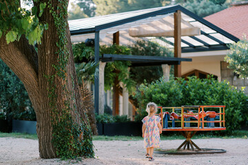 Little girl goes to the carousel in the playground. Back view