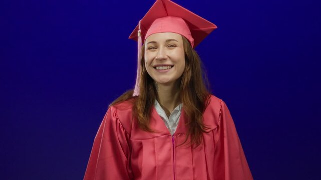 Young woman celebrating graduation in pink gown over blue wall pointing and smiling confidently.