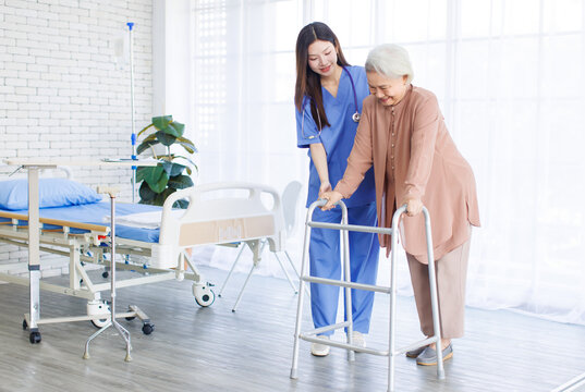 Asian doctor or caregiver helping assistance old woman to standing with walker in hospital, Young nurse supporting senior patient walk slowly frame nursing home care, Healthy wellness being concept.