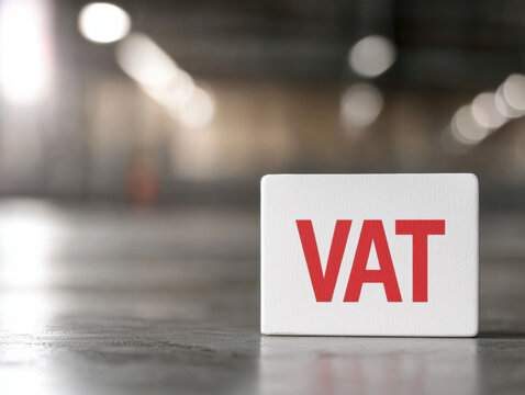 VAT displayed prominently in bold red font on white background, symbolizing tax concepts in modern, industrial setting. blurred background adds depth and focus to message