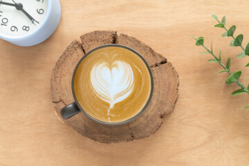 Concept for a banner, cafe, or coffee menu. A cappuccino latte art in the form of a heart in a dark brown ceramic cup, and a clock showing time 7:00, with green leaves on a rustic background..