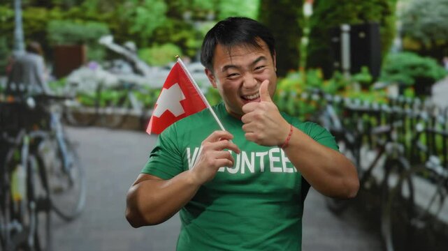 Young asian man holding switzerland flag standing outdoors in urban street showing positive expression in green volunteer shirt