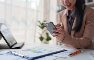 Smiling businesswoman using smartphone and analyzing financial charts, sitting at her desk with tablet and documents, working on business projects in modern office