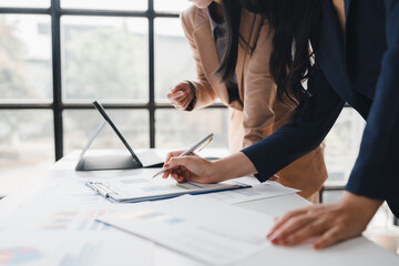 Two businesswomen are collaborating on a project, analyzing financial charts and data in a brightly lit office setting, using a tablet and paperwork