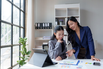Two asian businesswomen are collaborating on a project, analyzing financial charts and data at a modern office desk, working together to achieve business success