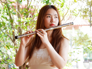 Young Asian woman playing a bamboo flute outdoors with green nature in the background, Traditional...