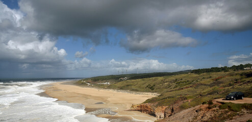 Ocean coastline with rolling white waves, yellow sandy beach, green grassy hills illuminated by midday sun, and rain clouds hanging low over landю Hilly landscape