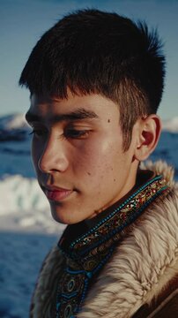 Young Inuit man with short black hair and a serious expression, wearing a traditional fur-lined coat with colorful patterns. Snowy landscape in Canada. Vertical shot