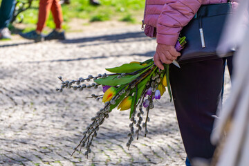 A person wearing a pink jacket holds a bouquet of flowers, which includes colorful tulips and willow branches (catkins). The photo was taken outside, on a cobblestone surface, on a sunny day © Adam