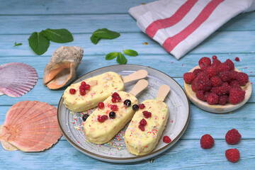 Homemade ice cream bars covered with white chocolate and red berries on a summery blue background, with raspberries and tropical decoration. Refreshing dessert concept