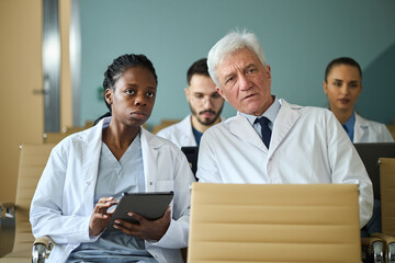 Group of healthcare professionals including an elderly senior discussing treatment strategy. Young male and female doctors focused on digital tablet showing serious expressions