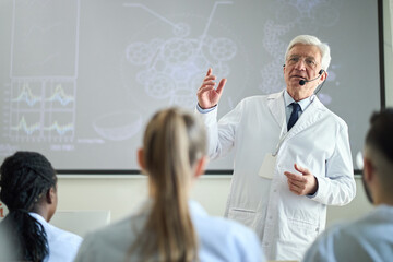 Elderly scientist explaining complex data during seminar while wearing lab coat. Diverse group of professionals attentively listening in classroom, enhancing medical knowledge