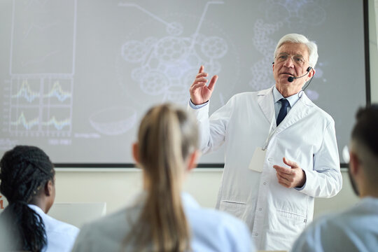 Elderly scientist explaining complex data during seminar while wearing lab coat. Diverse group of professionals attentively listening in classroom, enhancing medical knowledge