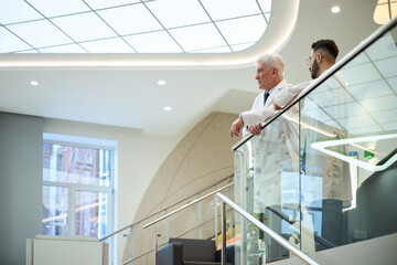 Two healthcare professionals, one elderly, standing in hospital discussing advancements. Captured in bright, modern environment with contemporary architectural features and large windows