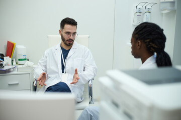 Two doctors engaging in a detailed conversation in medical office. Medical equipment and documents are visible, indicating a clinical setting and professional discussion