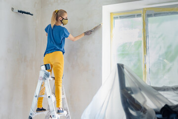 Young woman on stepladder cleaning wall after plastering works. Girl in a respirator sweeping a wall during apartment renovation, emphasizing solo repair work