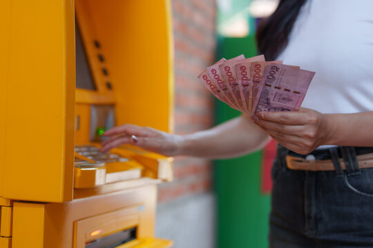 Woman withdrawing cash from an atm, holding thai baht banknotes in her hand, managing personal finances and utilizing modern banking services in a vibrant outdoor setting
