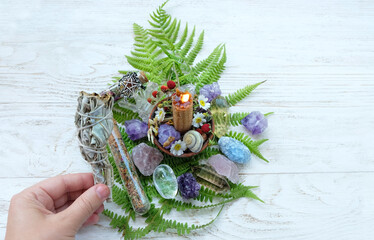 hand with smudge stick of white sage bundle, candle, minerals, flowers, leaves, amulet on table...