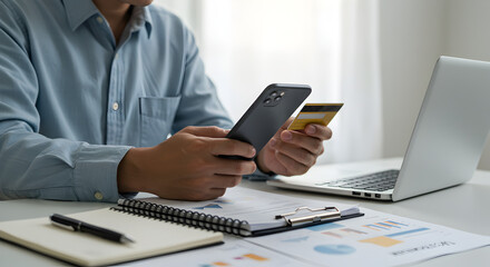 Fototapeta premium Person Holding Smartphone and Credit Card with Laptop and Charts on White Desk Showing Online Payment Process