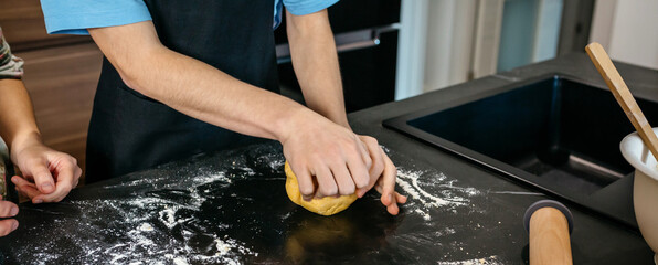 Banner of unrecognizable teenage boy with apron kneading dough while receives guidance from his mother, surrounded by kitchen counter filled with flour. Copy space available in image.
