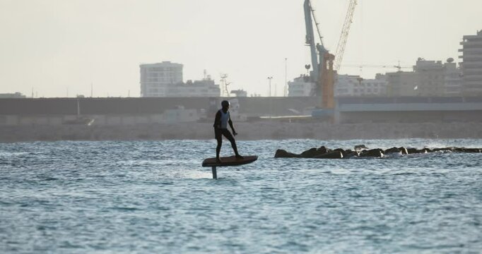A foilboard rider coasts across the sea as buildings rise behind in soft sunset haze. The mix of nature and city feels cinematic and atmospheric.. symbolizing liberation and solitude on the ocean surf