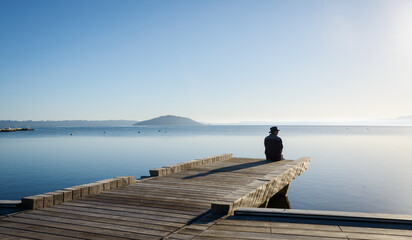 Man sitting on the boardwalk at lakefront of Lake Rotorua. Mokoia Island in the distance. Rotorua.