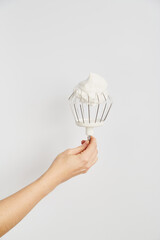 Hand holding a mixing whisk topped with whipped cream against a simple white background