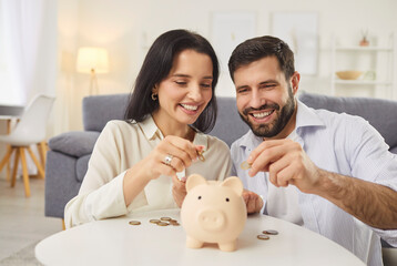 Family couple saving up some money for a mortgage or a car purchase. Happy smiling young married man and woman putting coins in a pink piggy bank on the table. Finance, budget, economy concepts