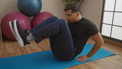 Young man exercising on a mat in a gym interior with exercise balls, showcasing his hispanic...