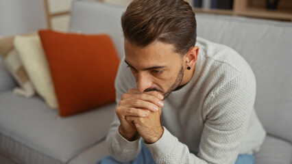 Obraz premium Young hispanic man indoors deep in thought at home sitting on a couch with a pensive look in a cozy living room setting surrounded by soft cushions.